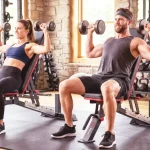 A dynamic photograph of an American man and woman engaged in strength training exercises using black adjustable weight benches in a well-equipped home gym.